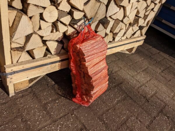 Het product, Aanmaakhout - Snel & Gemakkelijk Uw Vuur Aan van Onshout.nl, is verpakt in een rode netzak, rustend tegen een grote stapel brandhout. De grond bestaat uit donkere stenen, met zonlicht dat schaduwen creëert over het gebied.