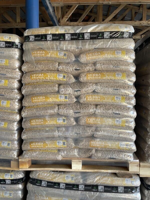 Stapels grote plastic zakken gevuld met Verba Premium 975kg ENplusA1 Houtpellets worden op houten pallets gelegd in een loods met zichtbare houten balken.