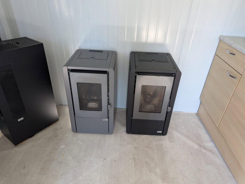 Twee slimme Tepor-pelletkachels van 7 kW met glazen deuren staan naast elkaar tegen een witte lambrisering, naast een zwart apparaat en een houten kast, op een lichte vloer. Beide zien er netjes gebruikt en goed onderhouden uit.