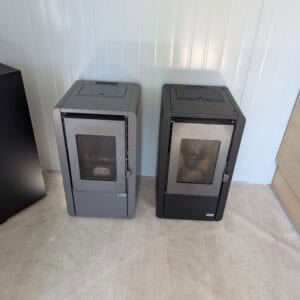 Twee slimme Tepor-pelletkachels van 7 kW met glazen deuren staan naast elkaar tegen een witte lambrisering, naast een zwart apparaat en een houten kast, op een lichte vloer. Beide zien er netjes gebruikt en goed onderhouden uit.