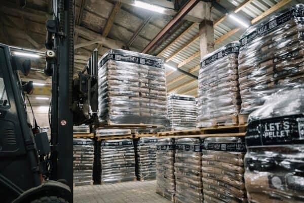 Een vorkheftruck tilt een omwikkelde pallet Let's Pellet 975kg ENplusA1 Houtpellets op in een magazijn vol gestapelde pallets met soortgelijke producten onder een hoog industrieel plafond.