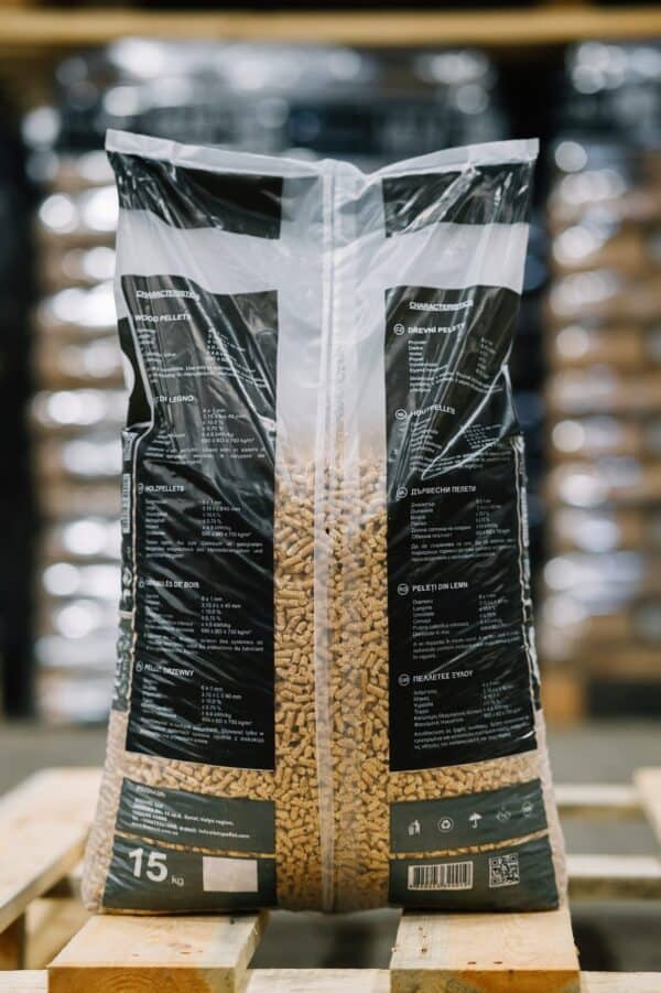 Een plastic zak van 15 kg Let's Pellet 975kg ENplusA1 Houtpellets rust op een houten pallet. De zak heeft zwarte en doorzichtige vakken, witte meertalige tekst en op de achtergrond zijn vage stapels pellets zichtbaar.