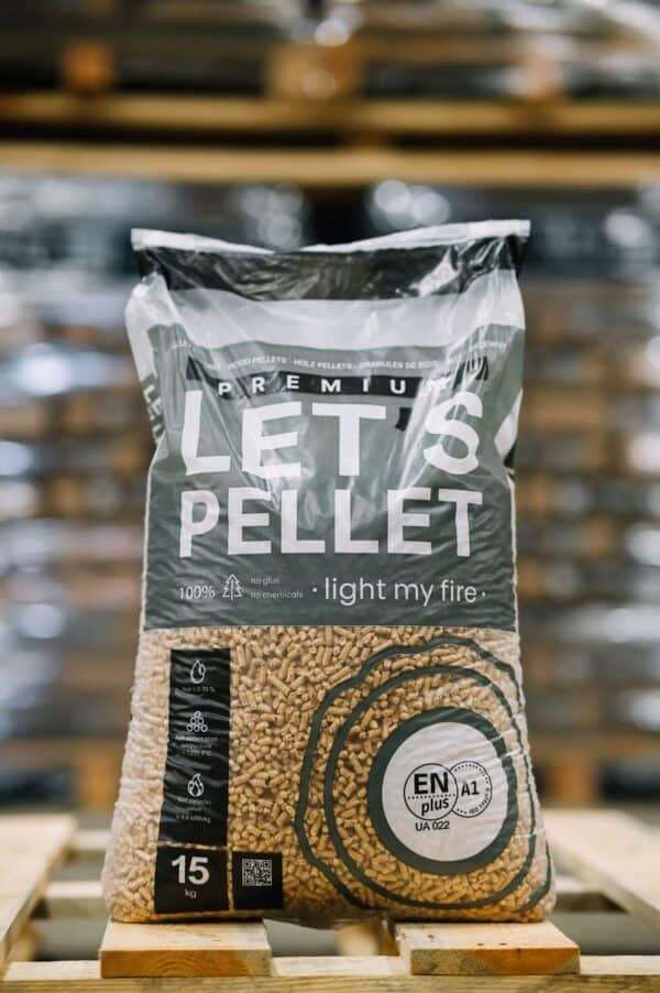 Een 15 kg Let's Pellet 975kg ENplusA1 Houtpellets zak met Verba Premium branding en certificeringspictogrammen wordt weergegeven op een "light my fire" pallet in een magazijnomgeving.